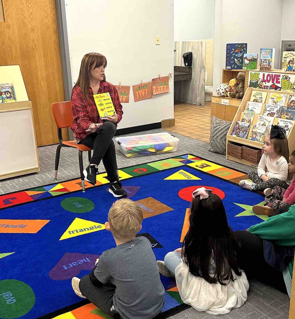 Rhonda sits on a chair on a blue carpet as she reads to a roomful of children.