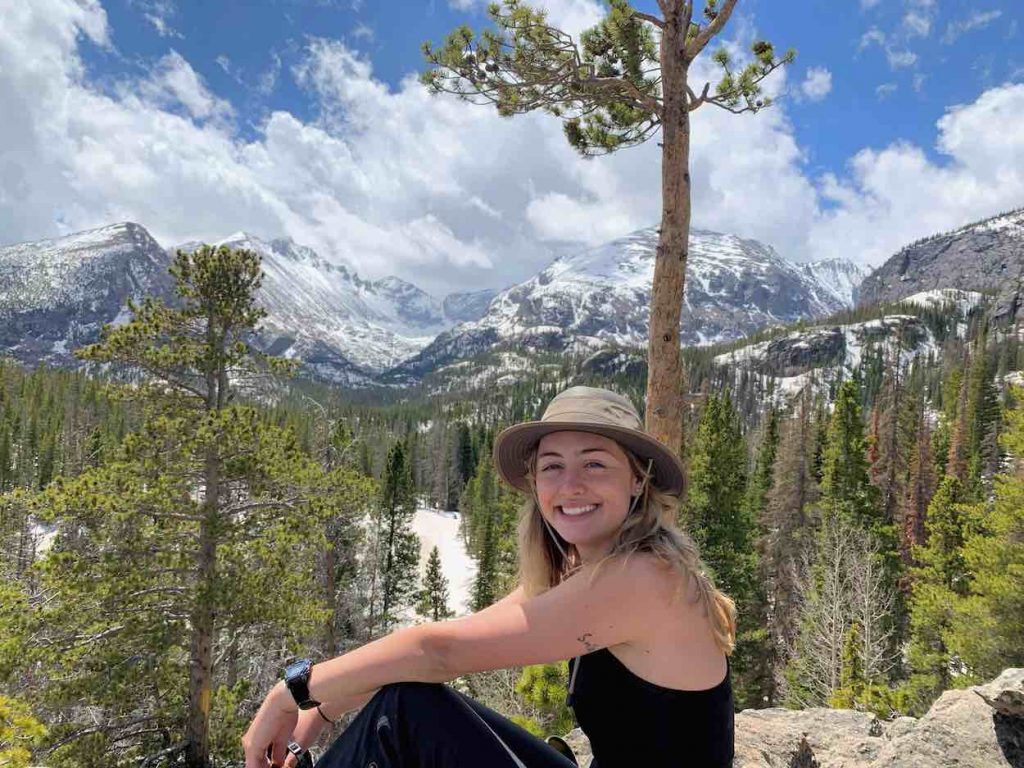Millie sits in a black tank top and pants with a khaki wide-brimmed hat with a mountain and blue skies in the background.