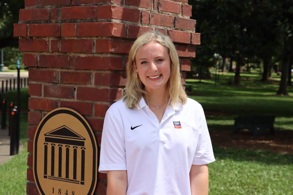 Emmaline stands in a white collared golf shirt with the SAA logo on it by the brick arch with the Ole Miss Lyceum. logo