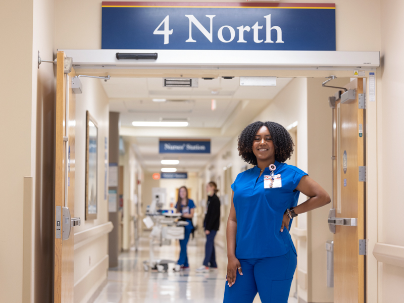 Tanzi McAllister wearing blue scrubs standing in a doorway under a sign that reads: 4 North