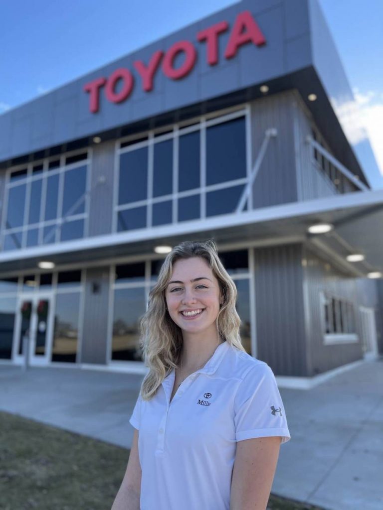 Millie stands in a white t-shirt in front of the Toyota plant. 