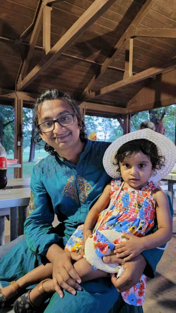 Khan wears a blue robe and holds his daughter who is a wearing a sun dress and a white straw hat. They are sitting outside under a pavilion.