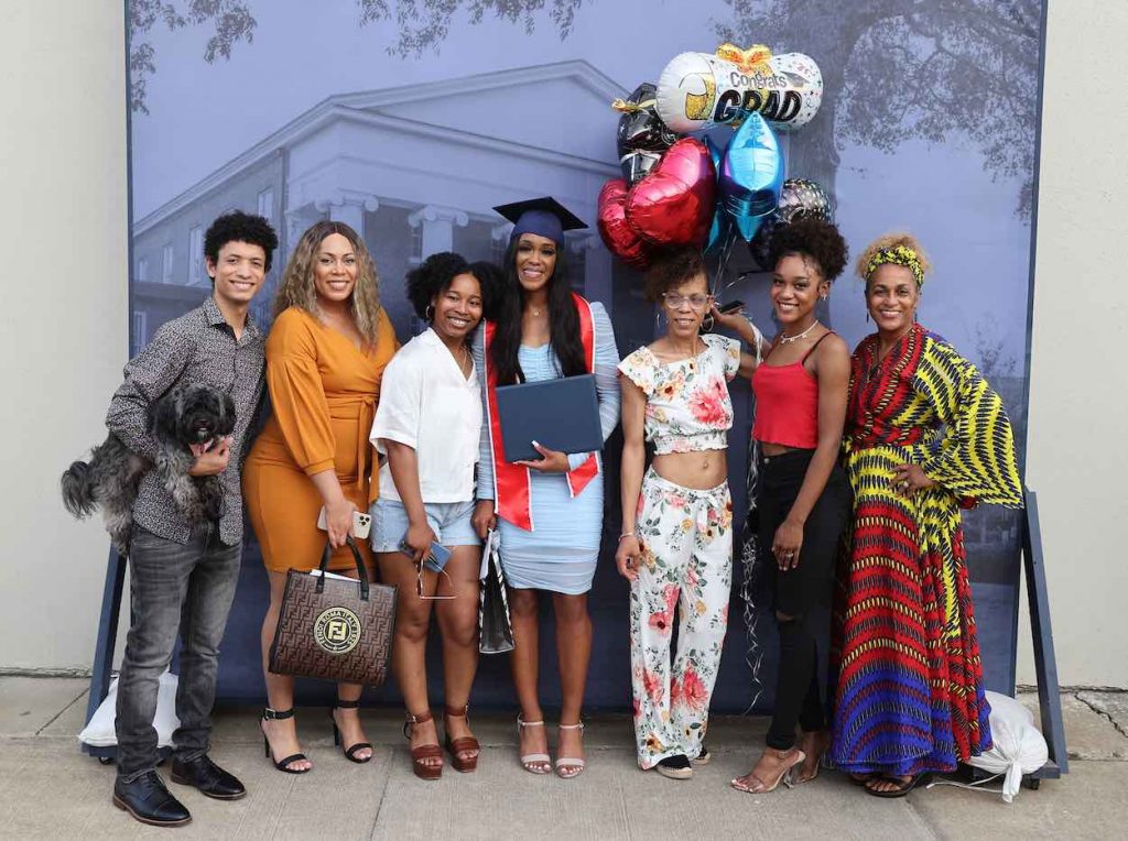 Orianna wears a powder blue dress in her navy blue graduation cap and red stole. She is standing with six friends in front of screen and balloons.
