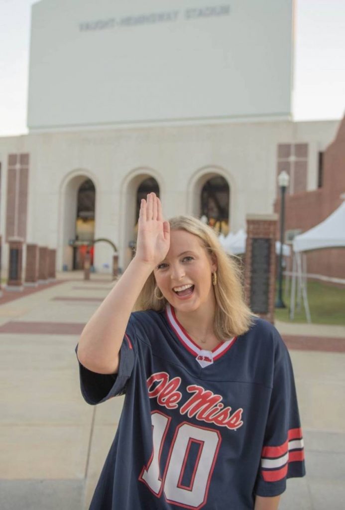 Emmaline does the Fins Up sign on her forward as she stands in a #10 Ole Miss football jersey in front of a football stadium.