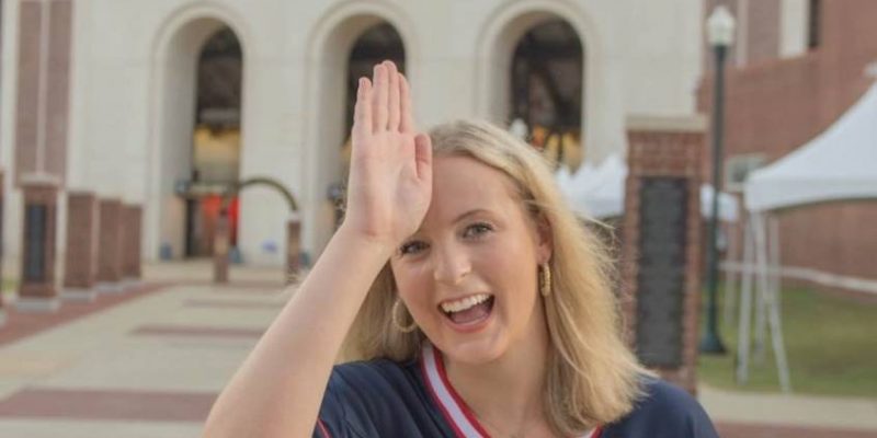 Emmaline does the Fins Up sign on her forward as she stands in a #10 Ole Miss football jersey in front of a football stadium.