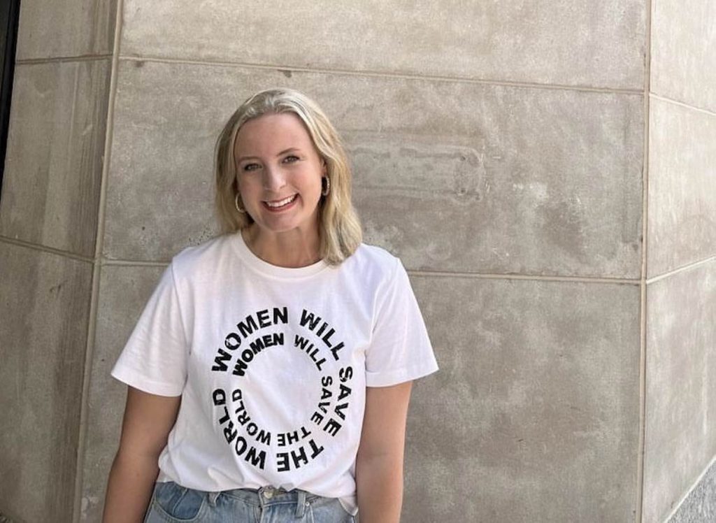 Emmaline stands in front of a white stone building in a white t-shirt that says, "Women will change the world."