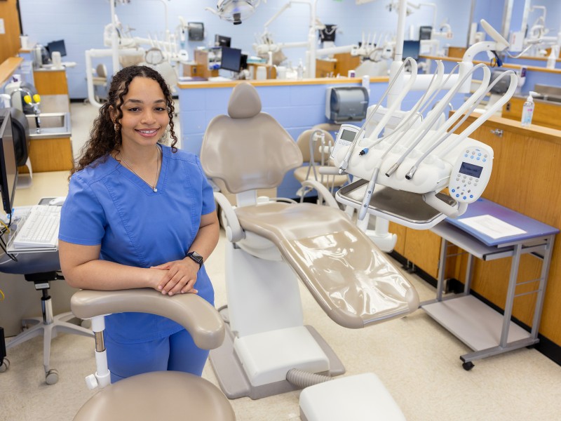 Malita Chaffier stands in blue scrubs in front of a dental chair and equipment in a medical office. 