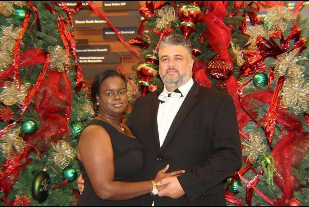 Bailey wears a black suit standing in front of red and green holiday decorations next to his wife who is wearing a black dress.